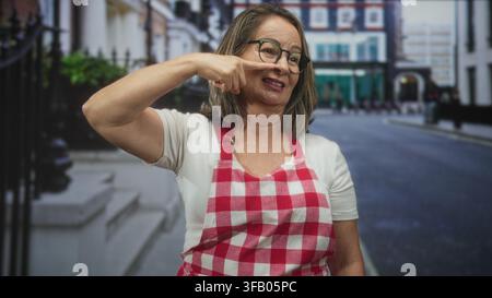 Die ranghöchste hispanische Frau zeigt Finger an Nase, während sie rote karierte Schürze und Brille auf der Stadtstraße in der Nähe der Treppen des Stadthauses trägt; verspielte Wärme. Stockfoto