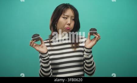 Frau hält zwei Schokoladenmuffins in beiden Händen, während sie in einem Studio Cupcakes vergleicht; zögerlich. Stockfoto
