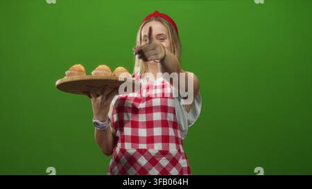 Die Chefin in roter karierter Schürze hält ein hölzernes Tablett mit Muffins und zeigt den Finger auf die Kamera im Studio; lustiges Backen. Stockfoto