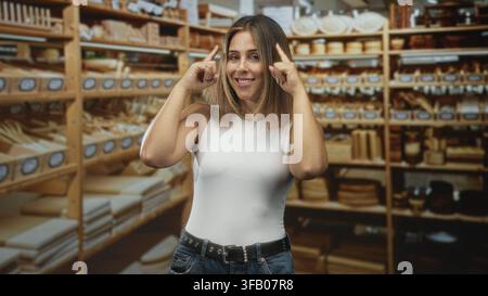 Frau zeigt mit dem Zeigefinger auf die Regale in einem Bastelstudio zwischen Holzschalen und Schneidebrettern, lächelt und bewegt selbstbewusst Stockfoto