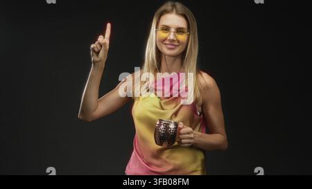 Die blonde Frau zeigt den Zeigefinger nach oben, während sie in einem Studio einen Kupferbecher hält. Stockfoto