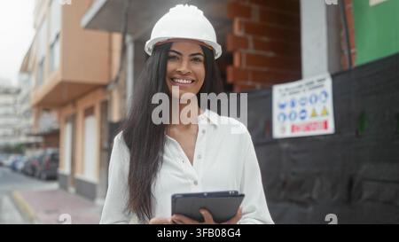 Frau hält ein Tablet und schaut lächelnd auf Pläne, trägt einen Helm und inspiziert eine Baustelle auf einem städtischen Bürgersteig; Selbstvertrauen le Stockfoto