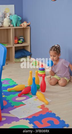 Mädchen hält einen blauen Ball, sitzt auf dem Boden und greift nach Plastik-Kegelspiel auf einem Schaumstoff-Puzzleteppich, gestapelten Ringen und Zeichnungen in einem Spielzimmer insi Stockfoto