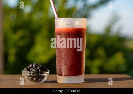 Frischer Blaubeer-Smoothie in einem hohen Glas im Freien, Nahaufnahme. Gekühlter dunkler Beeren-Smoothie oder Saft in einem mattierten Glas mit Strohhalm neben frischen Blaubeeren o Stockfoto