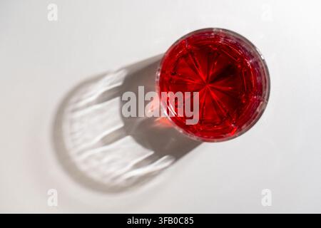 Top-Down-Ansicht von rotem Getränk in einem Kristallglas mit schönem Schatten, Nahaufnahme. Hochwinkelaufnahme eines facettierten Glases, gefüllt mit lebhafter roter Flüssigkeit, das Schatten wirft Stockfoto