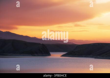 Ein ruhiger Canyon-Fluss oder See, flankiert von dunklen Hügeln unter einem leuchtend orangefarbenen und gelben Sonnenuntergangshimmel mit geschichteten Bergkämmen in der Ferne. Ideal f Stockfoto