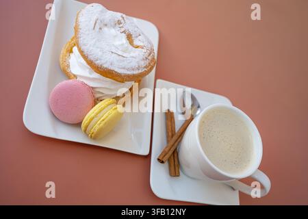 Vertikales ästhetisches Arrangement von heißem Cappuccino in einer Keramiktasse neben bunten Macarons, zarter Baiser und dekorativen Zimtstangen auf dem Tisch Stockfoto