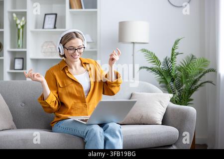 Fröhliche junge Frau mit Kopfhörern, die auf einem gemütlichen Sofa lächelt und tanzt, während sie zu Hause Musik hört oder Inhalte auf ihrem Laptop streamt, entspannt und unbeschwert Stockfoto