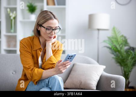 Frau mit Brille sitzt auf einem bequemen Sofa zu Hause, vertieft in ihr Smartphone, um mit Menschen in Kontakt zu treten, in sozialen Medien zu surfen oder aus der Ferne zu arbeiten Stockfoto