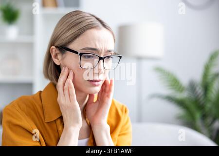Reife Frau mit Brille, die ihre Wange berührt und vor starker Zahn- oder Kiefer Schmerzen zusammenzuckt, gestresst und unwohl drinnen in einer Zahnarztpraxis oder bei einem Arzttermin, Nahaufnahme Stockfoto