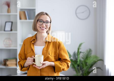 Junge Frau mit Brille lächelt, genießt eine warme Tasse während einer entspannten Pause in ihrem hellen Homeoffice, strahlt Komfort, Selbstvertrauen und gemütliche, produktive Stimmung aus Stockfoto