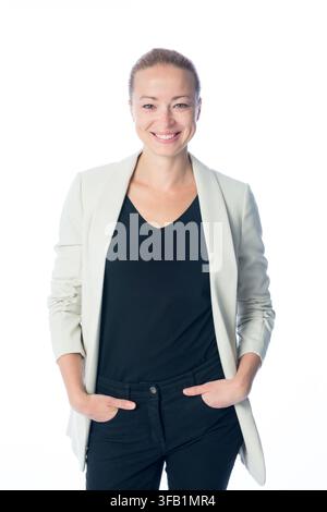 Studienporträt einer fröhlichen blonden Geschäftsfrau in einem hellgrauen Blazer und einem schwarzen Oberteil, die selbstbewusst lächelt, während ihre Hände in den Taschen stecken, vor einem neutralen weißen Hintergrund Stockfoto