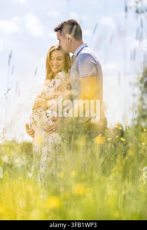 Zärtliches Hochformat-Porträt eines Ehemannes, der die Stirn seiner lächelnden schwangeren Frau küsst, während sie sich umarmen und ihren Babybauch halten, stehend in einem üppigen Wildblumenfeld Stockfoto