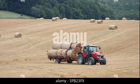 Bardejov, Slowakei – 19. August 2026: Ein roter Traktor zieht einen Anhänger, der mit runden Heuballen beladen ist, über ein abgeerntetes Feld, fotografiert im Somme Stockfoto