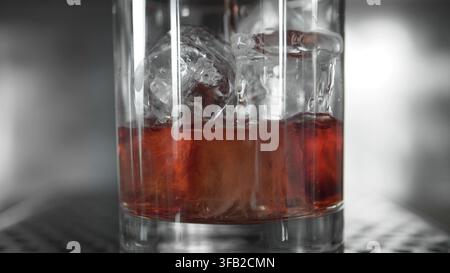 Braune Flüssigkeit, die in ein mit Eis gefülltes Glas gegossen wird, erzeugt Spritzer und Blasen. Zubereitung eines erfrischenden alkoholischen Getränks Stockfoto