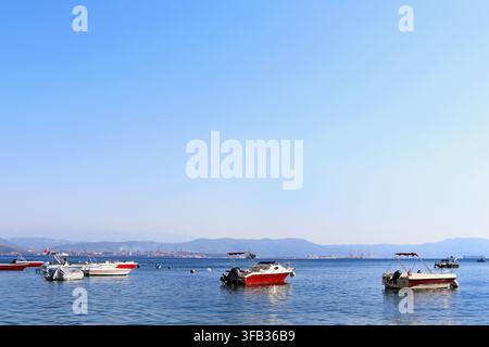 Boote, die in einer ruhigen Bucht mit Bergen, Frachtschiffen und einem Industriehafen am Horizont vor Anker liegen Stockfoto