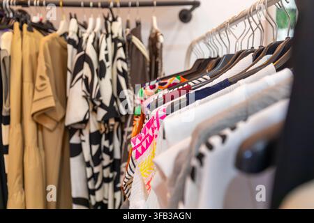 Nahaufnahme von farbenfroher Modekleidung, die auf Kleiderständern in einer Boutique hängt, mit gemusterten Kleidern, Hemden und neutraler Kleidung in einer Stockfoto