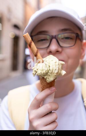 Lächelnder Teenager mit Brille und Kappe hält eine Pistazieneiswaffel mit Waffelstock, während er eine historische Straße in Rom, Italien, im Sommer erkundet Stockfoto