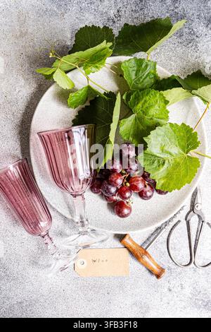 Überkopf-Stillleben mit reifen roten Trauben, Weinrebenblättern, rosa Weingläsern und einer Astschere, die auf einem Keramikteller über einer strukturierten grauen Oberfläche angeordnet sind Stockfoto