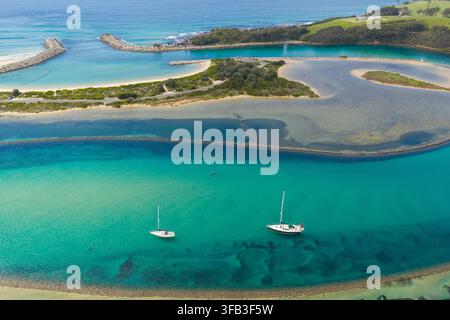 Luftaufnahme von türkisfarbenen Buchteinwässern in Narooma, New South Wales, Australien, mit zwei vor Anker liegenden Segelbooten, sandigen Kanälen, Wellenbrechern, Küstenvegetation Stockfoto