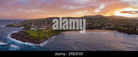 Luftbild von Kendalls Beach bei Sonnenuntergang, das die geschwungene Küstenlinie, Küstenhäuser, grünes Kap und rollende Brandung in Kiama, New South Wales, Australien zeigt Stockfoto