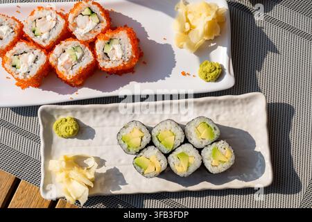 Nahaufnahme von verschiedenen Sushi-Rollen mit Reis, Gurke, Avocado, Ingwer, cremiger Füllung und leuchtendem Lachsrogen, angeordnet auf weißen Keramiktellern über einem Stockfoto