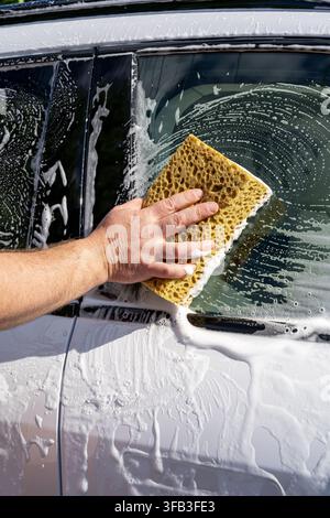 Nahaufnahme einer Hand, die ein Autofenster im Freien mit einem gelben Schwamm, dickem Seifenschaum und Wassertröpfchen während der Fahrzeugreinigung und -wartung wäscht Stockfoto