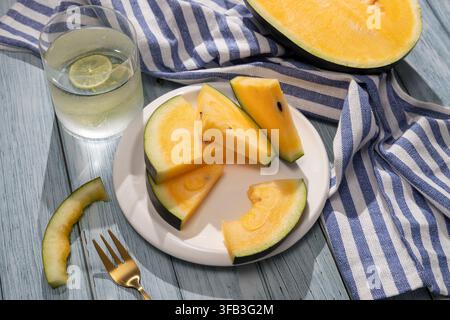Ansicht von oben auf gelbe Wassermelonenstücke auf einem weißen Teller neben einem gekühlten Limettengetränk, gestreifter Stoff und Gabel auf einem rustikalen blauen Holztisch. Stockfoto