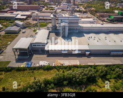 Hohe Luftaufnahme zeigt große weiße Produktionsanlage mit Industrietürmen neben einem Lagerbereich, der mit Waren und grüner Vegetation gefüllt ist, unter Stockfoto