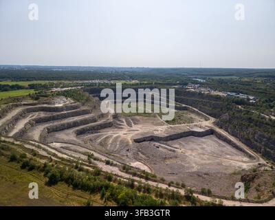 Hochwinkelaufnahme zeigt eine massive terrassierte Granitabbaustätte mit gewundenen unbefestigten Transportwegen neben grünem Wald und landwirtschaftlichen Wegen Stockfoto