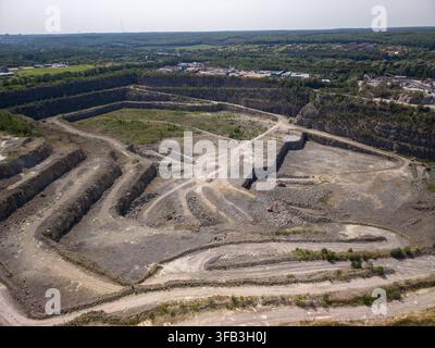 Hohe Drohnenaufnahme zeigt ein großes offenes Steinbruch mit felsigen Terrassen, Schotterspuren, Kieshaufen und arbeitenden Baggern in der Nähe Stockfoto