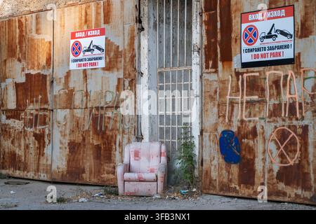 Ein verlassener, verwitterter Sessel steht auf der Straße vor stark verrosteten Industrietüren, die mit verblasstem Graffiti und einem "No Parking"-Schild gekennzeichnet sind Stockfoto