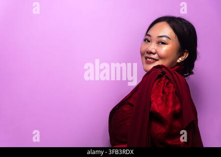 Lächelnde junge asiatische Frau in einem kastanienbraunen Outfit, die die Kamera auf lila Hintergrund mit Kopierfläche ansieht Stockfoto