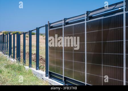 ein moderner Metallzaun, der mit integrierten vertikalen Solarpanelen konstruiert wurde, entlang einer Betonbasis in einem trockenen, offenen Feld unter einem klaren blauen Himmel platziert ist. Stockfoto