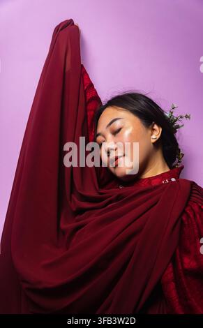 Künstlerisches Porträt einer schönen jungen asiatischen Frau, die einen kastanienbraunen Vorhang vor lila Hintergrund hält Stockfoto