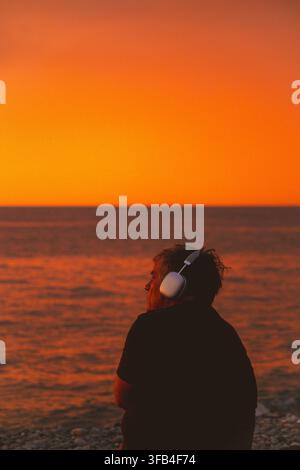 Ein Mann beobachtet den Sonnenuntergang vor der Kulisse des Meeres, während er Kopfhörer trägt Stockfoto