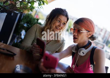 Zwei Freunde sitzen zusammen in einem Café in einer Stadt, lächeln und schauen auf ein Handy, teilen einen Moment am Tag Stockfoto