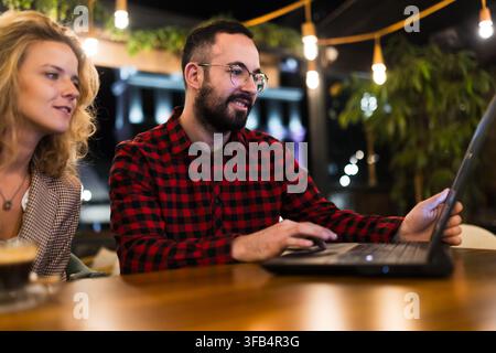 Junger Mann und Frau arbeiten an einem Laptop in einem gemütlichen Café mit warmer Beleuchtung und einer freundlichen Atmosphäre, tragen lässige Kleidung und genießen eine produktive Zeit Stockfoto