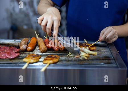 Eine Nahaufnahme eines Straßenhändler, der mit Metallzangen eine Vielzahl von Fleischstücken, Würsten und Satay-Spießen über einem kommerziellen Gasgrill wendet, foo Stockfoto