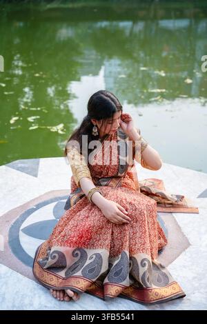 Eine Frau in einem traditionellen Saree mit aufwendigen Mustern sitzt an einem Gewässer und hält ein Mobiltelefon ans Ohr. Stockfoto