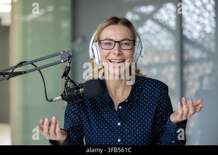 Glückliche junge Frau mit Brille und Kopfhörern spricht in ein Mikrofon in einem modernen Studio, lächelt während der Aufnahme eines Podcasts oder einer Live-Übertragung für Online-Medien und Streaming Stockfoto