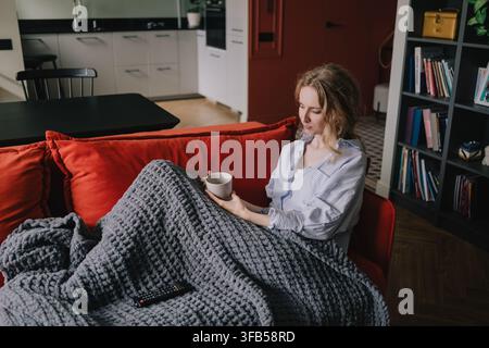 Frau ruht unter einer warmen Decke mit einer Tasse, während sie zu Hause auf einem Sofa entspannt Stockfoto