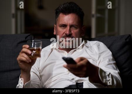Porträt eines traurigen Mannes mittleren Alters, der Whiskey und eine TV-Fernbedienung hält Stockfoto