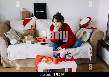 Frau in gestricktem Pullover arbeitet an einem Laptop auf dem Sofa neben verpackten Paketen und einem HOLLY JOLLY-Schild. Remote-Arbeit Dezember, Laptop-Heimbüro Weihnachten Stockfoto