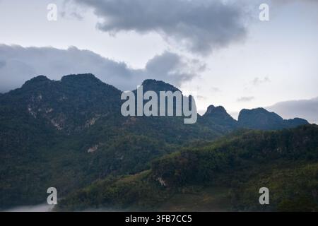 Neblige Kalksteinberge, die mit üppigem grünen Wald bedeckt sind, unter einem dramatischen bewölkten Himmel, der eine friedliche tropische Landschaft mit schroffen Gipfeln und Nebel schafft Stockfoto