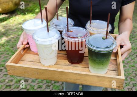 Ein Holztablett mit bunten Eistränken und Strohhalmen im Freien, Person trägt verschiedene Getränke während eines lockeren Erfrischungsmoments in einer Gartensituation Stockfoto