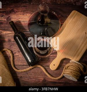 Stillleben mit Rotwein: großes Glasdekanter, gefülltes Weinglas, unbeschriftete Flasche und altertöniger Tonkrug, arrangiert mit Sackleinen und Seil auf rustikalem Holz Stockfoto