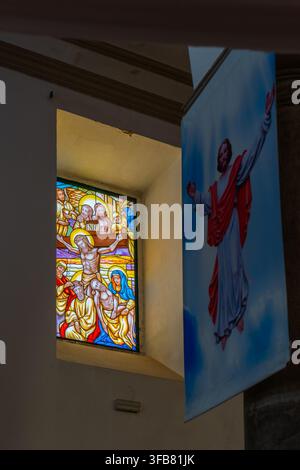 Helle Kreuzigungsszene aus Buntglas erscheint in einer Kirchennische neben einem hängenden Banner, das Jesus darstellt. Stockfoto