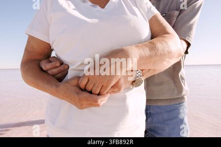 Nahaufnahme eines älteren Paares, das sich mit verschlungenen Händen am Pink Salt Lake umarmt. lebenslange Liebe, Vertrauen, emotionale Unterstützung, Hingabe, Ehe Stockfoto