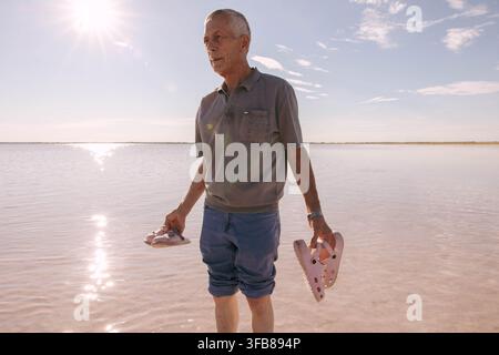 Senior mit flachem rosa Salzwasser, der in beiden Händen rosa Kinderschuhe hält. Helles Sonnenlicht, ruhige Wasseroberfläche, blauer Himmel mit Licht Stockfoto